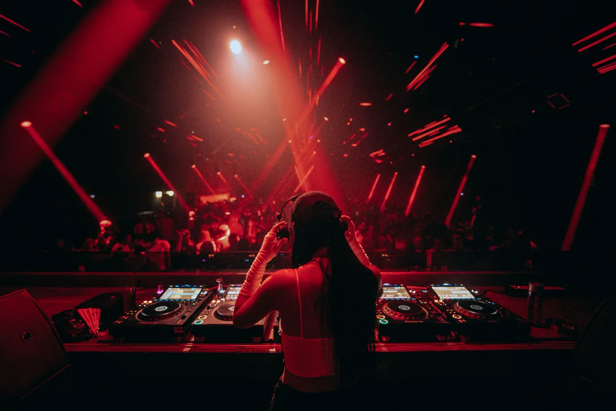DJ silhouetted at the decks, facing a dark crowd under red beams of light